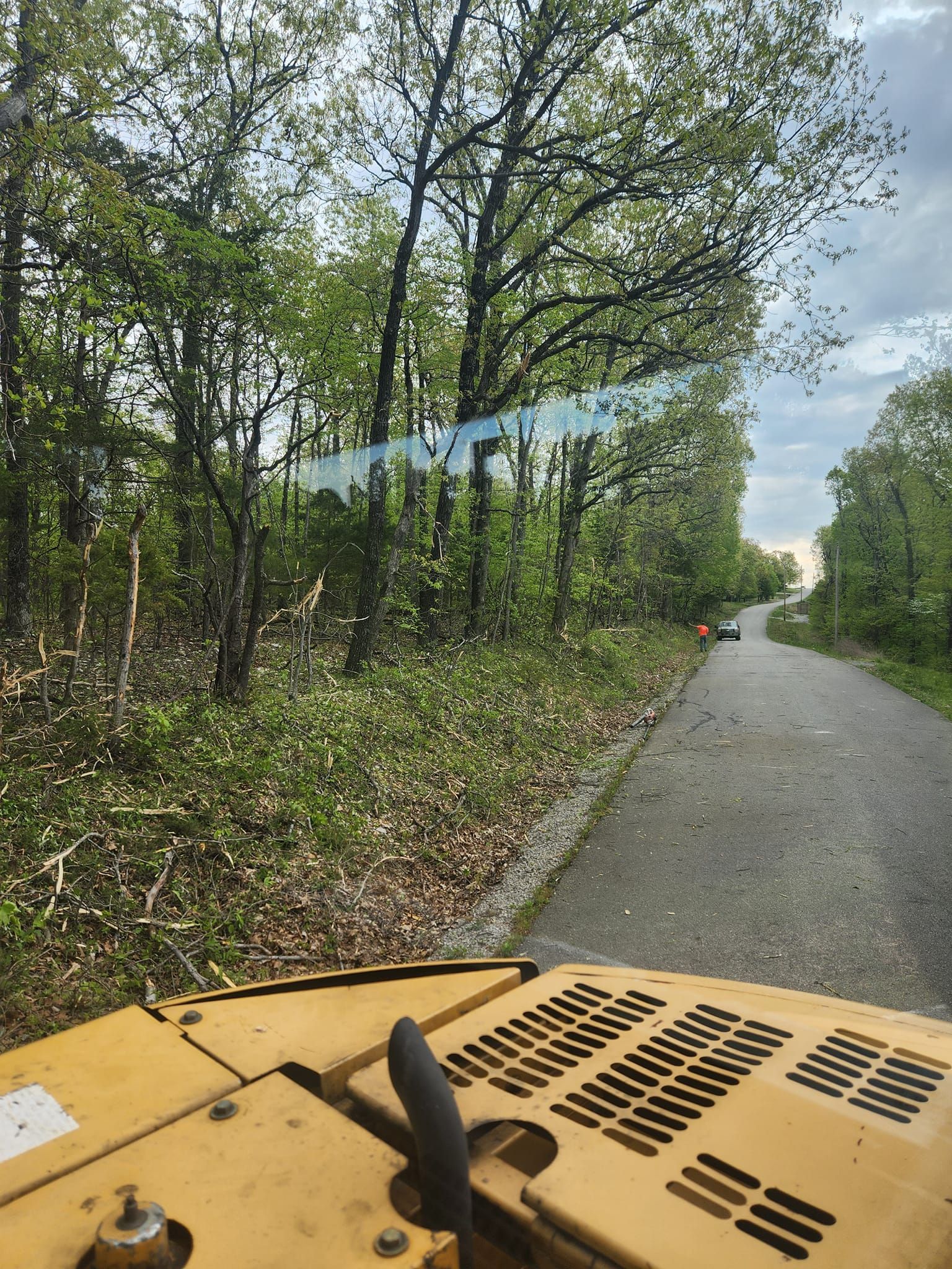 A person operating heavy machinery on a gravel path alongside a wooded area with lush green foliage.