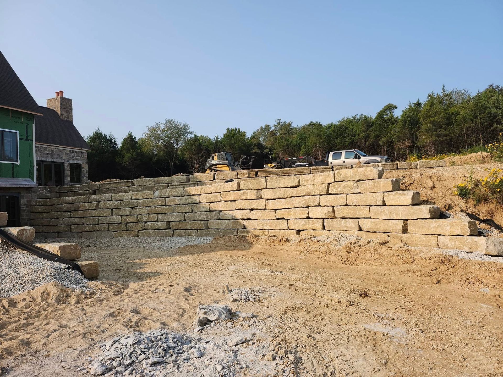 A large retaining wall made of light-colored stone is under construction next to a house and a hillside.