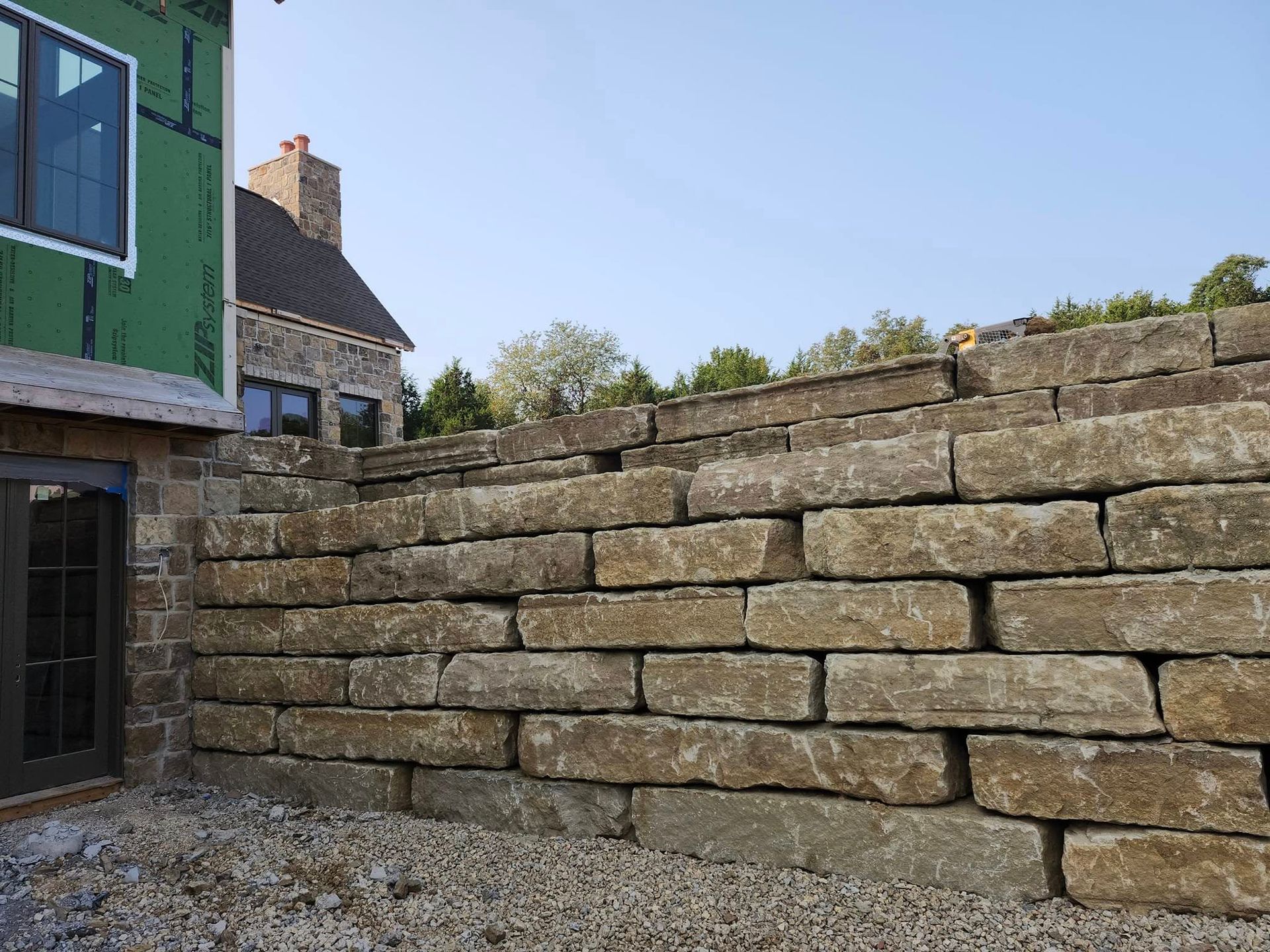 A stone retaining wall in front of a house. The wall is made of light brown rectangular stones stacked in rows.