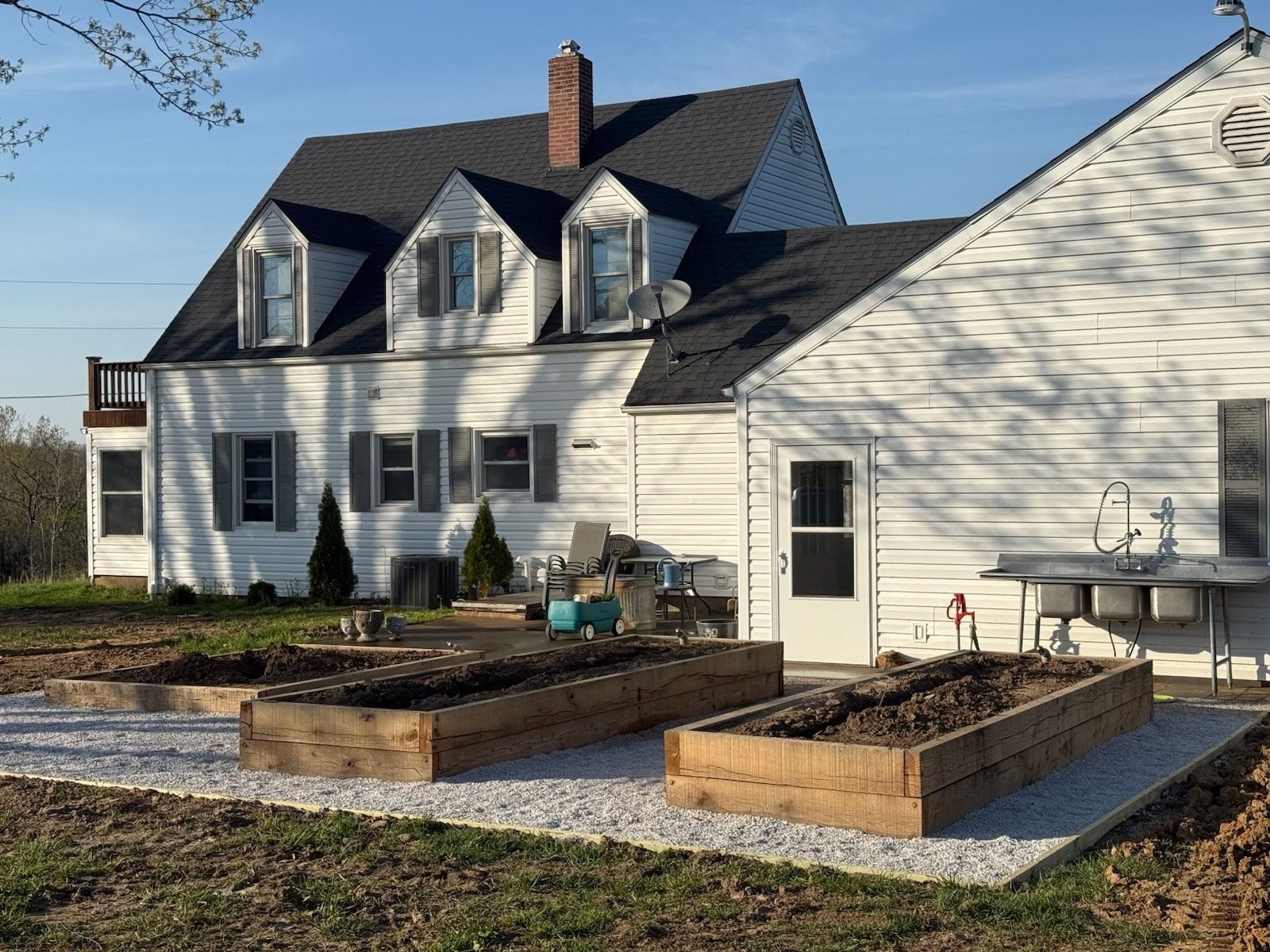 Raised garden beds in front of a white house with a dark roof. A gravel pathway surrounds the beds.