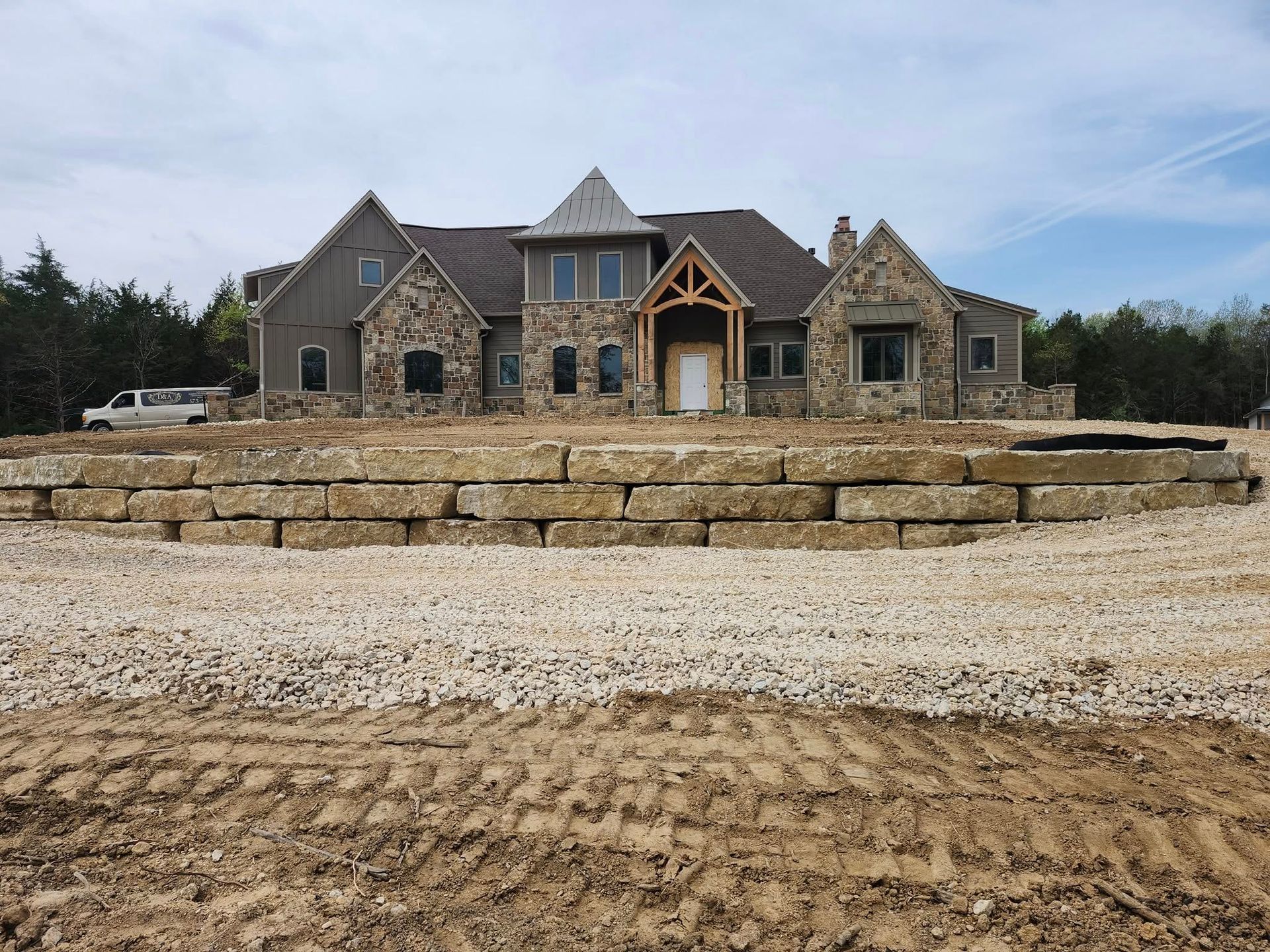 A large stone house under construction with a gravel driveway and a retaining wall made of stacked stone blocks.