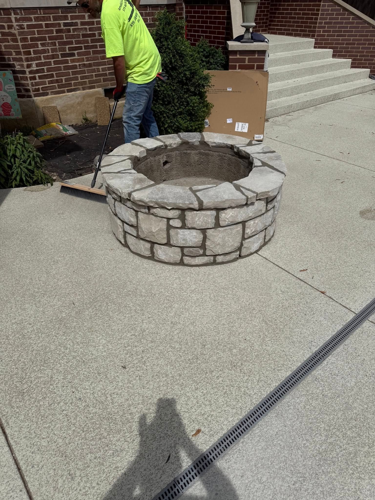 A stone fire pit on a concrete surface with a person in a yellow shirt working nearby.