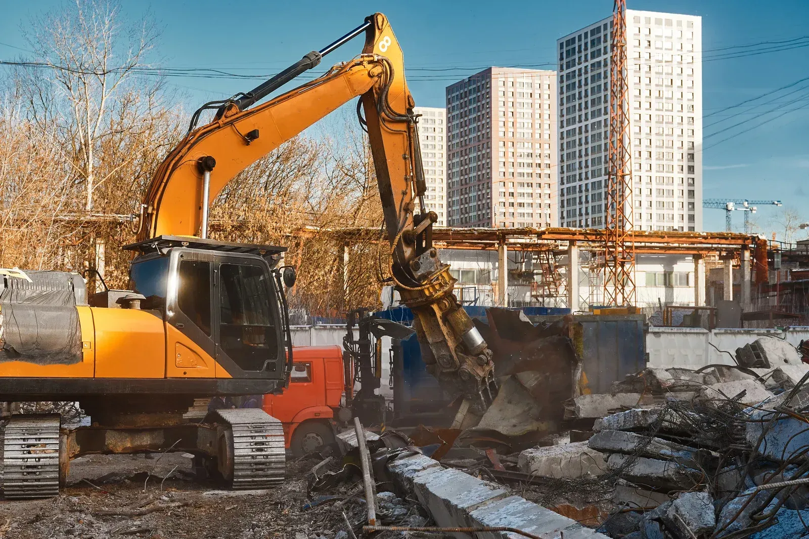 An orange excavator demolishes concrete structures in front of several high-rise buildings under a clear sky.