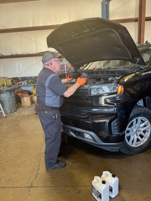 Mechanic working on a black truck with hood open in a garage. Two fluid containers are on the floor.