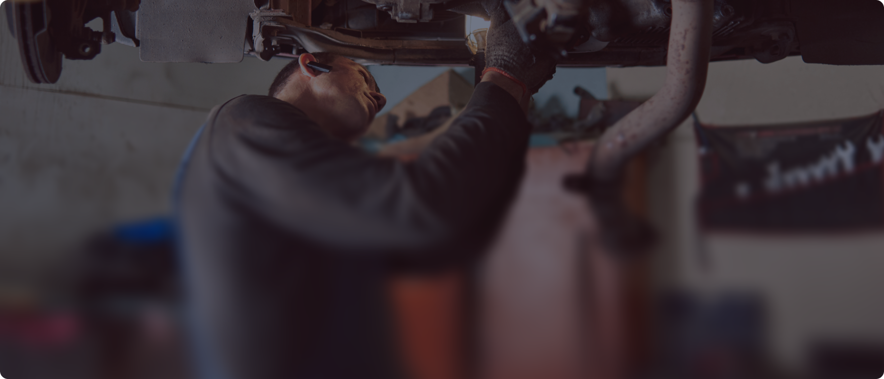 A person working under a vehicle. They are wearing a long-sleeve shirt and gloves. The setting is a garage.