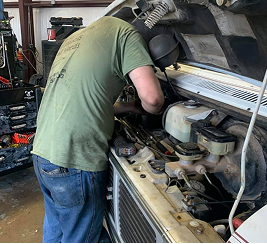 Mechanic working on the engine of a vehicle with the hood open. Wearing a green t-shirt, jeans, inside a garage.