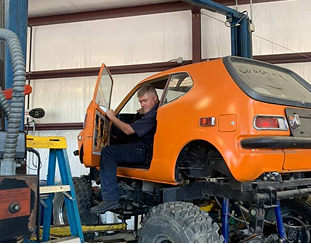 Man in orange car raised on monster truck tires in a garage.