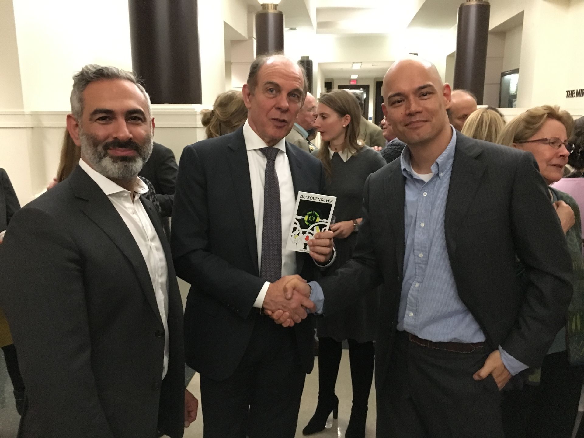 Three people in business attire shake hands in a hallway, with the person in the middle holding a small white booklet.