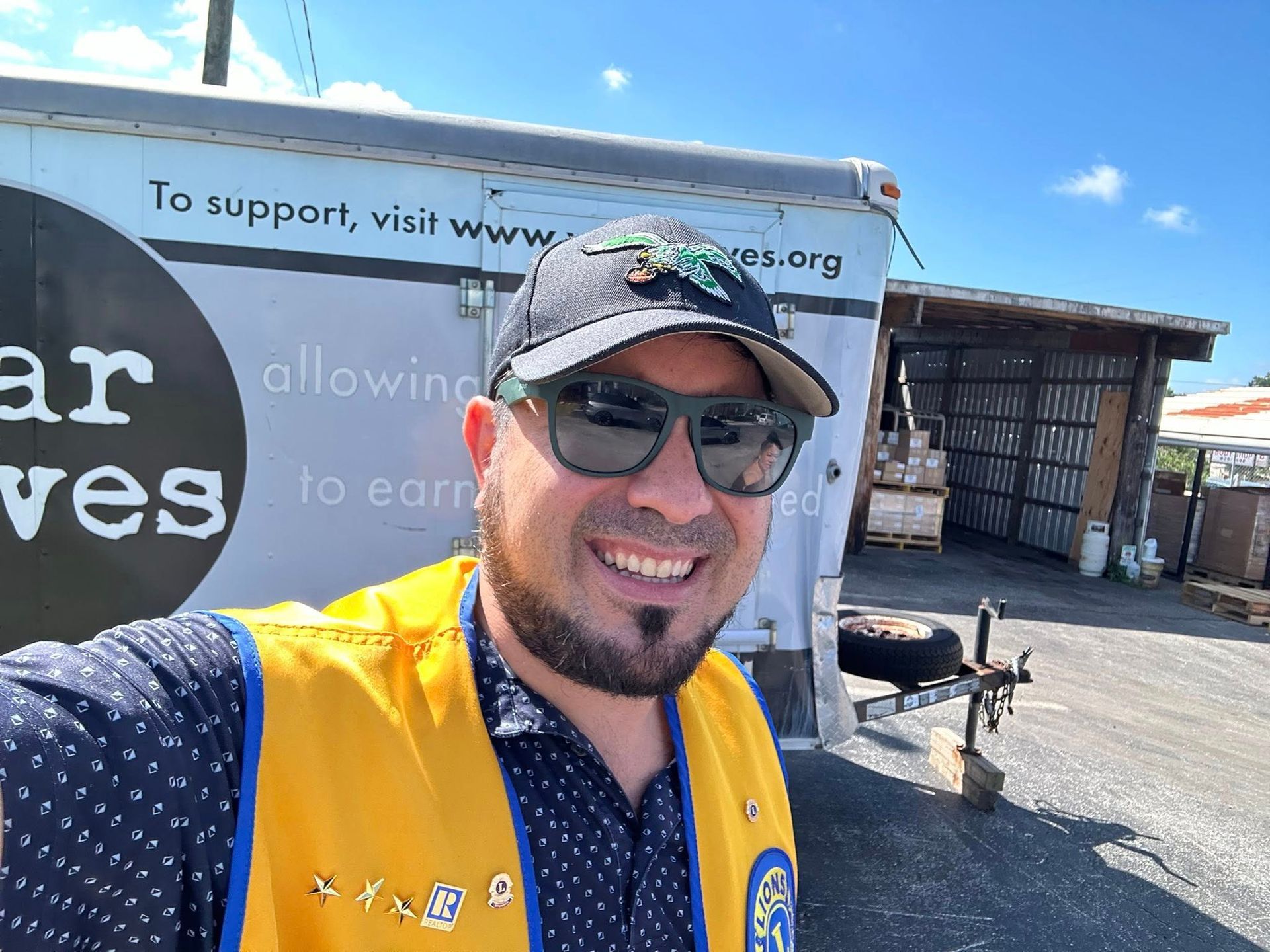 Man in sunglasses, baseball cap, and yellow vest smiles in front of a trailer on a sunny day.