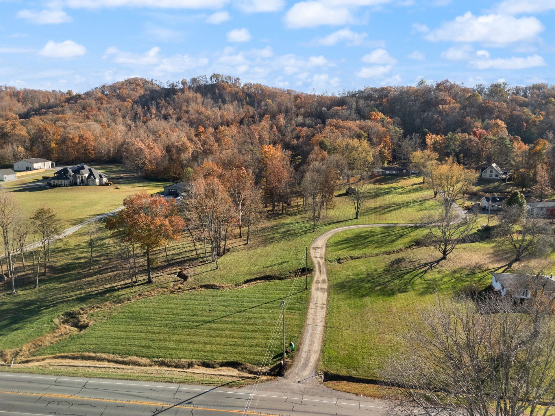 Dirt road winds uphill through a grassy field towards a tree-covered hillside under a partly cloudy sky.