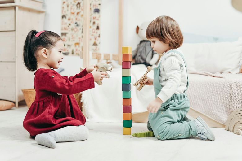Two children playing with blocks and toys in a bedroom, separated by a column of blocks.