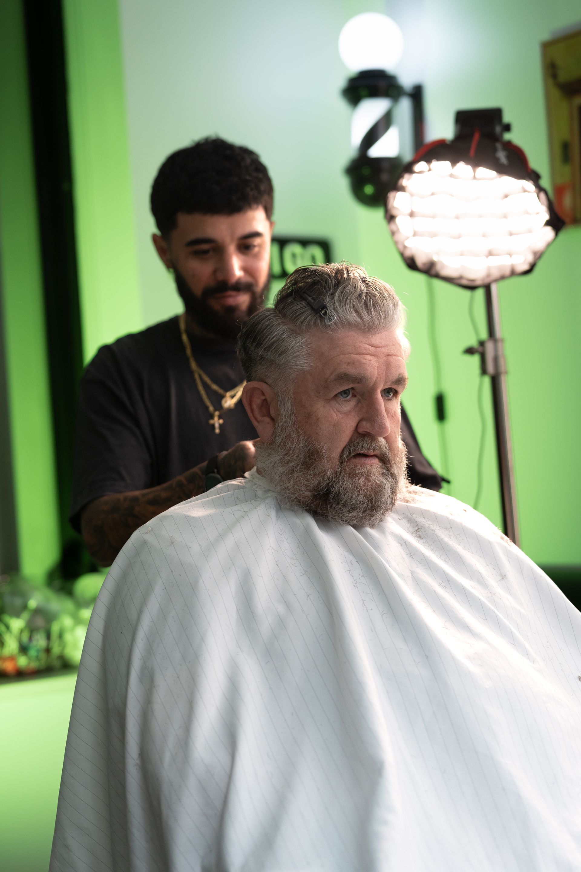Barber giving a haircut to a client in a barbershop with a green-tinted wall and bright lights.