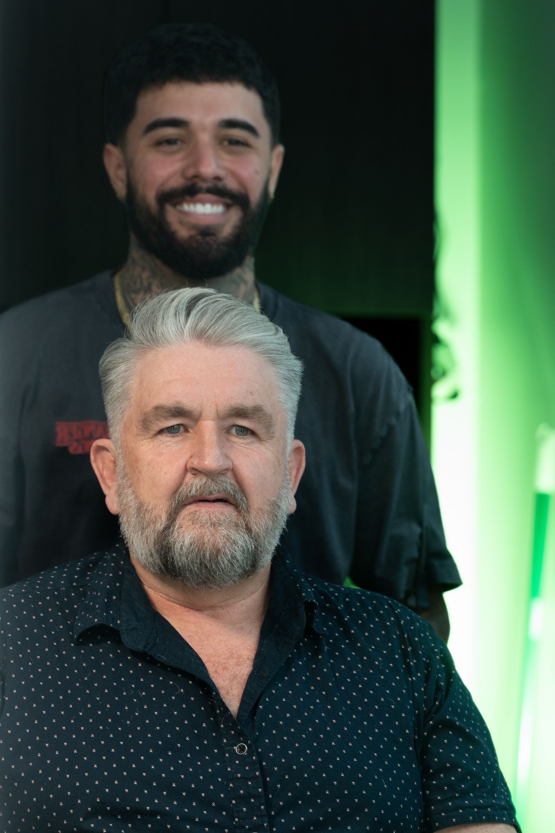 Two men posing together: older with gray beard, younger with tattoos and smile, green background.
