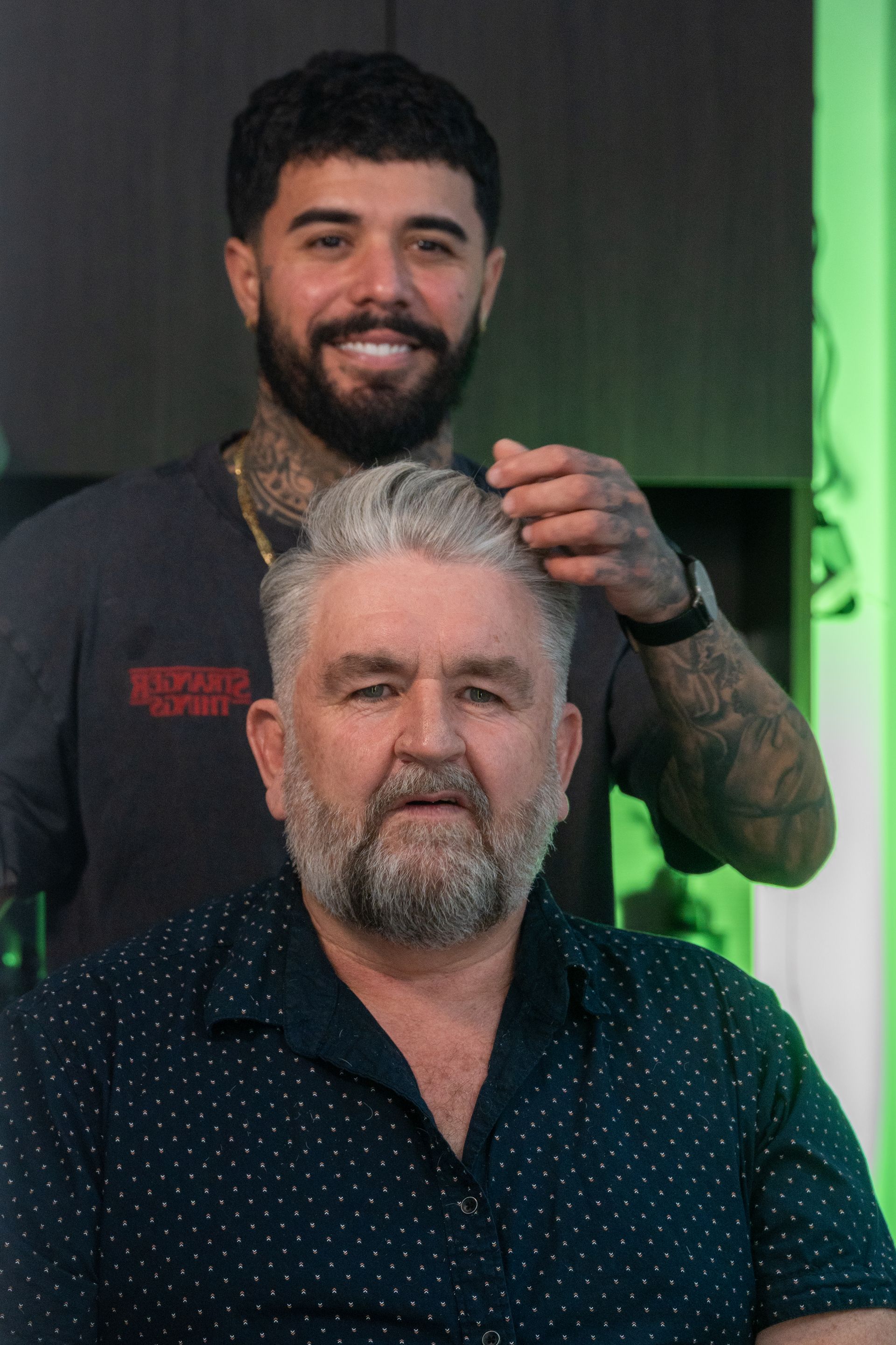 Barber adjusting client's hair in a barber shop. Both are smiling. Green-lit background.