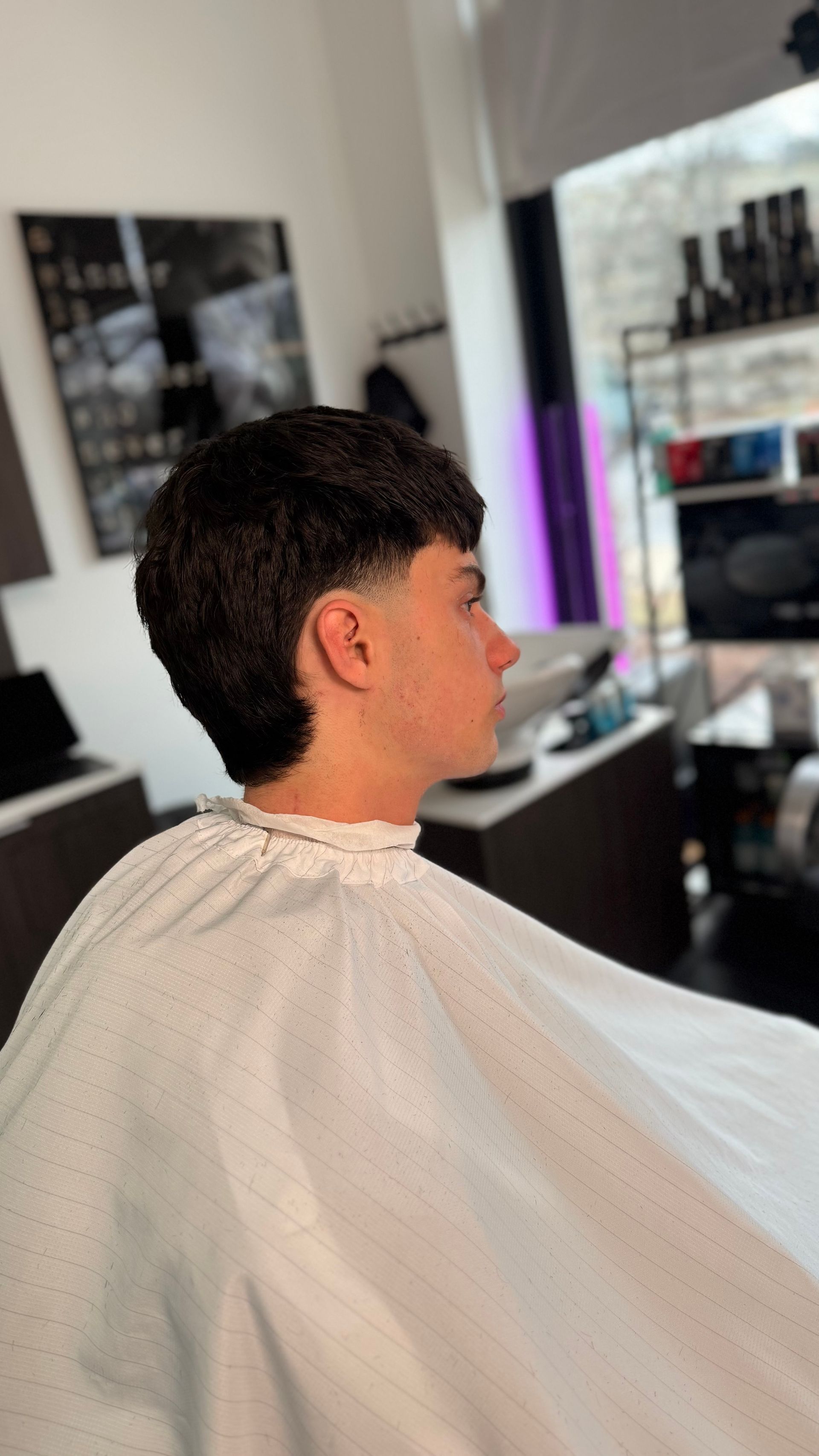 Person with a dark haircut in a barber shop, wearing a white cape. Side view.