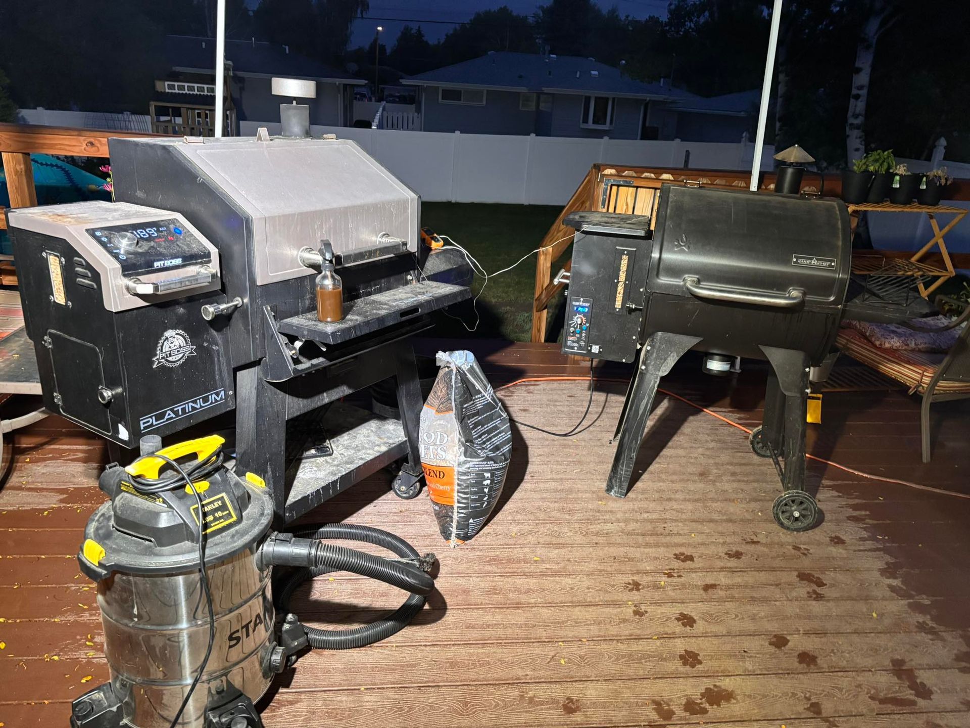 Two BBQ smokers on a wooden deck, with a shop vac in front. Nighttime setting.
