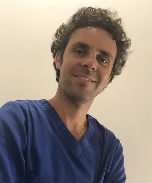 Un uomo con i capelli scuri e ricci sorride, indossando una camicetta blu, in un ambiente ben illuminato.