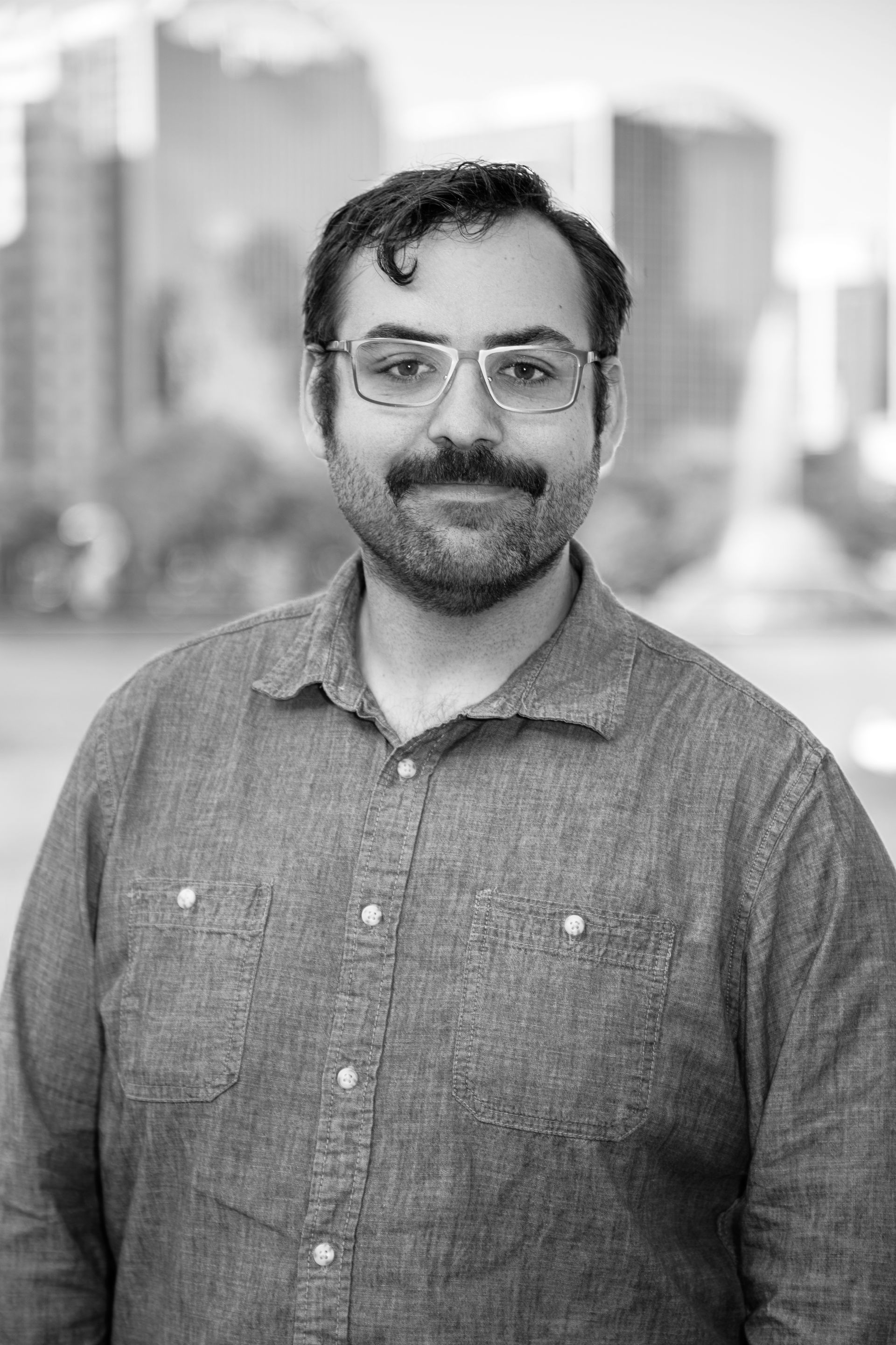 A man with a beard and glasses is standing in front of a city skyline in a black and white photo.