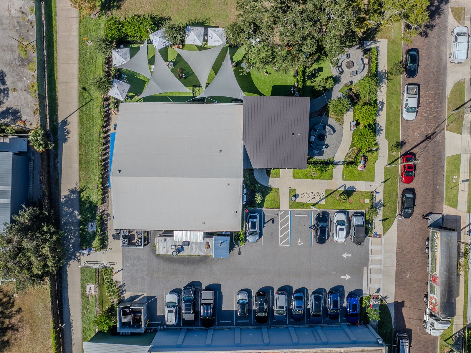 Aerial view of a building with a parking lot and a landscaped outdoor area with shade sails. Cars parked on the street.