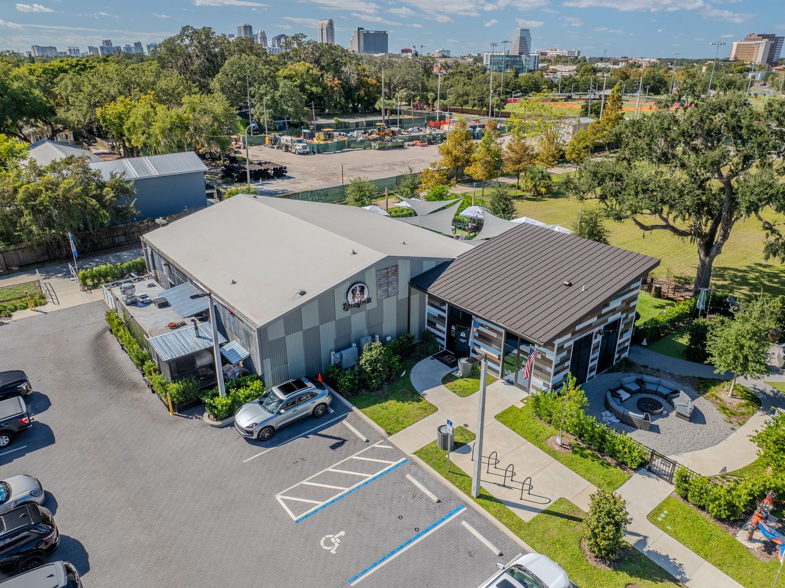 Aerial view of a modern building with corrugated metal siding, a patio, and parking lot, surrounded by greenery.
