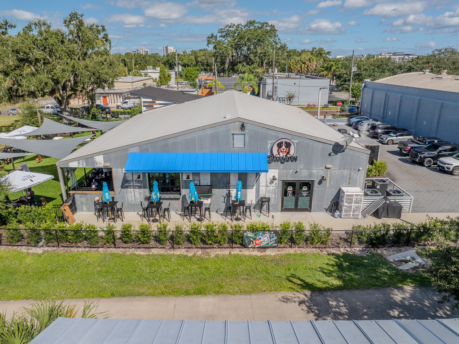 Restaurant with a blue awning and outdoor seating, set against a backdrop of trees and parked cars.