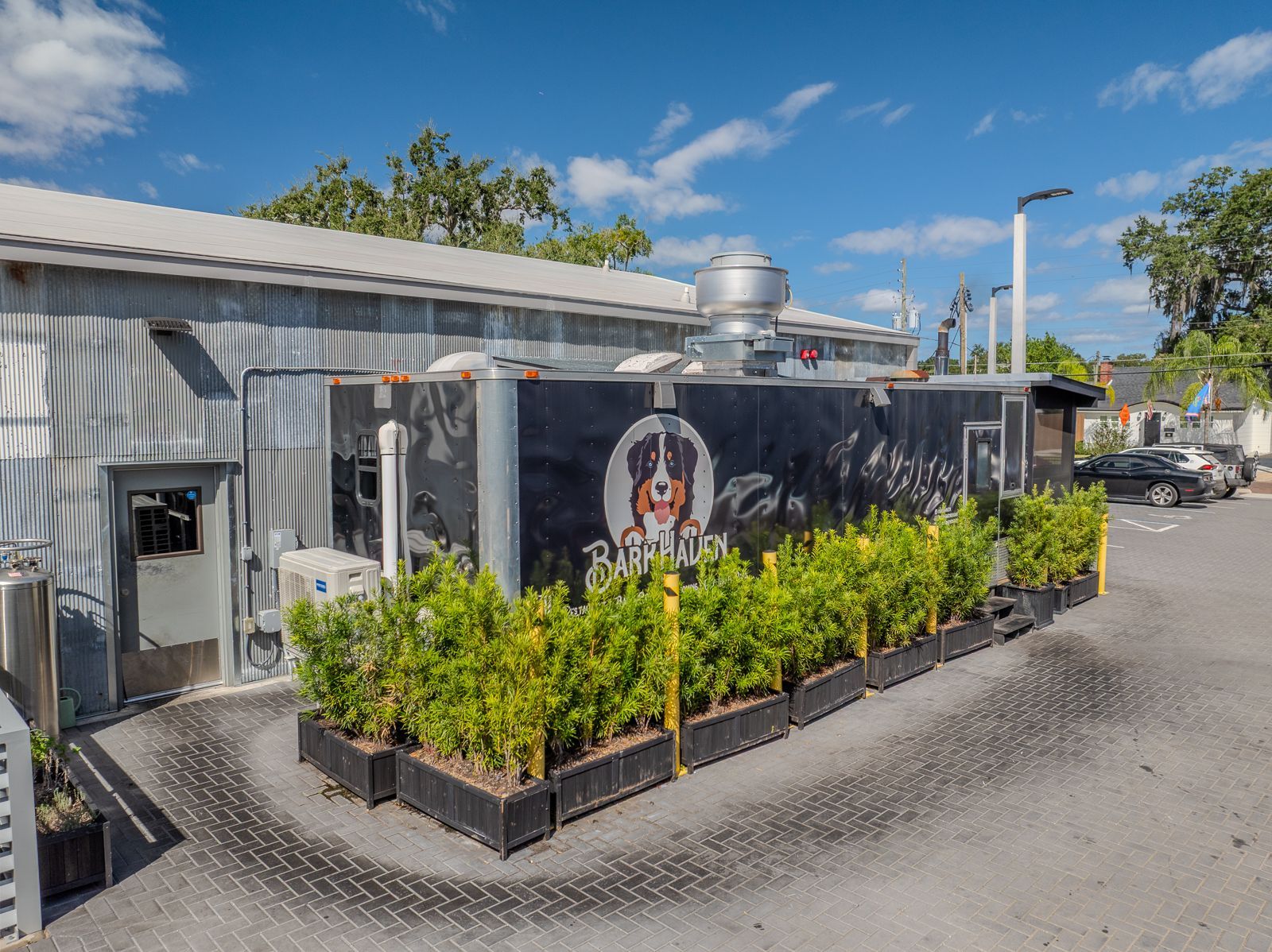 Restaurant exterior with black and corrugated metal walls. Green plants line the sidewalk. Blue sky.