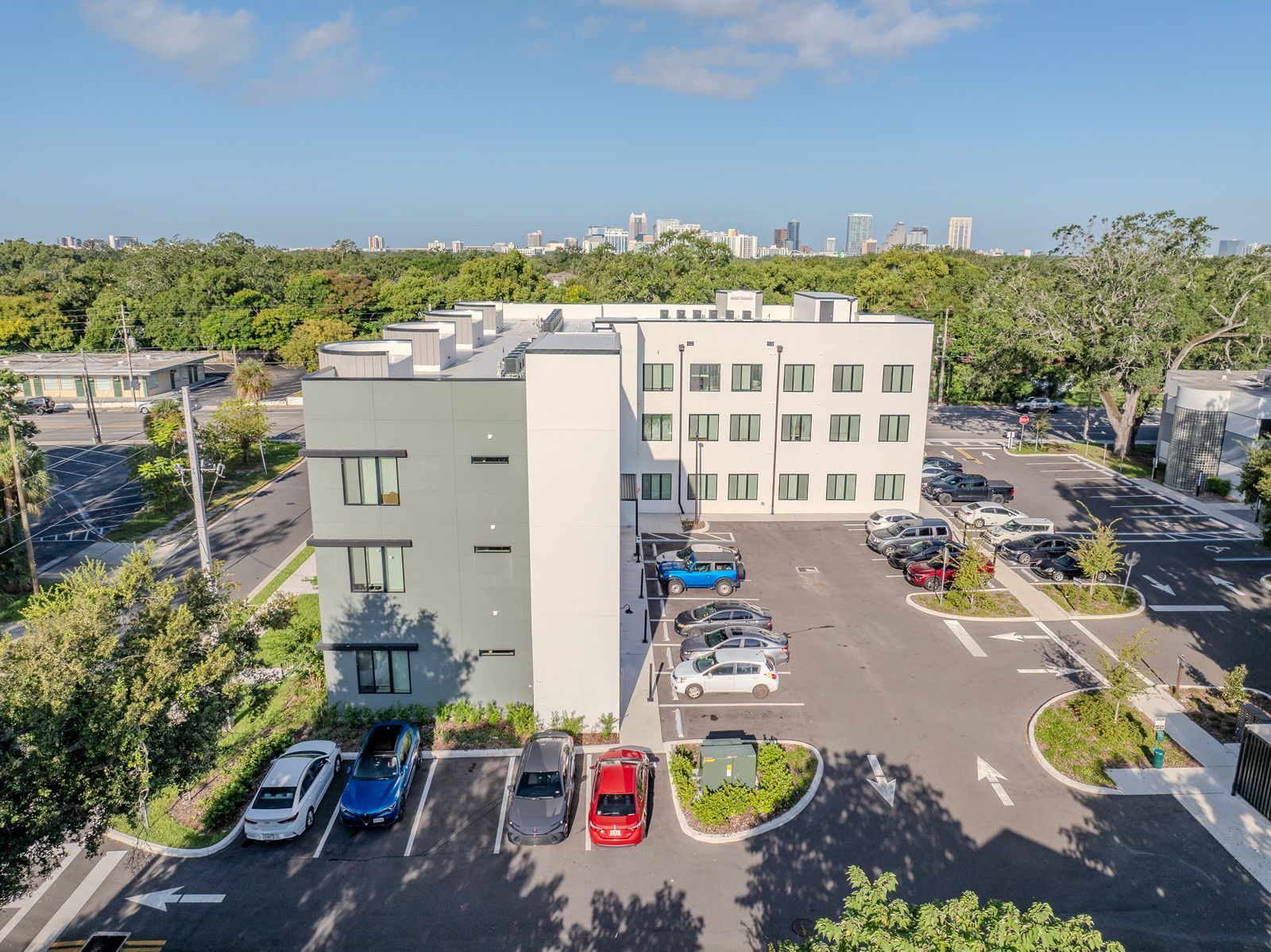 Modern multi-story building with a parking lot and cars, trees and a city skyline in the distance.