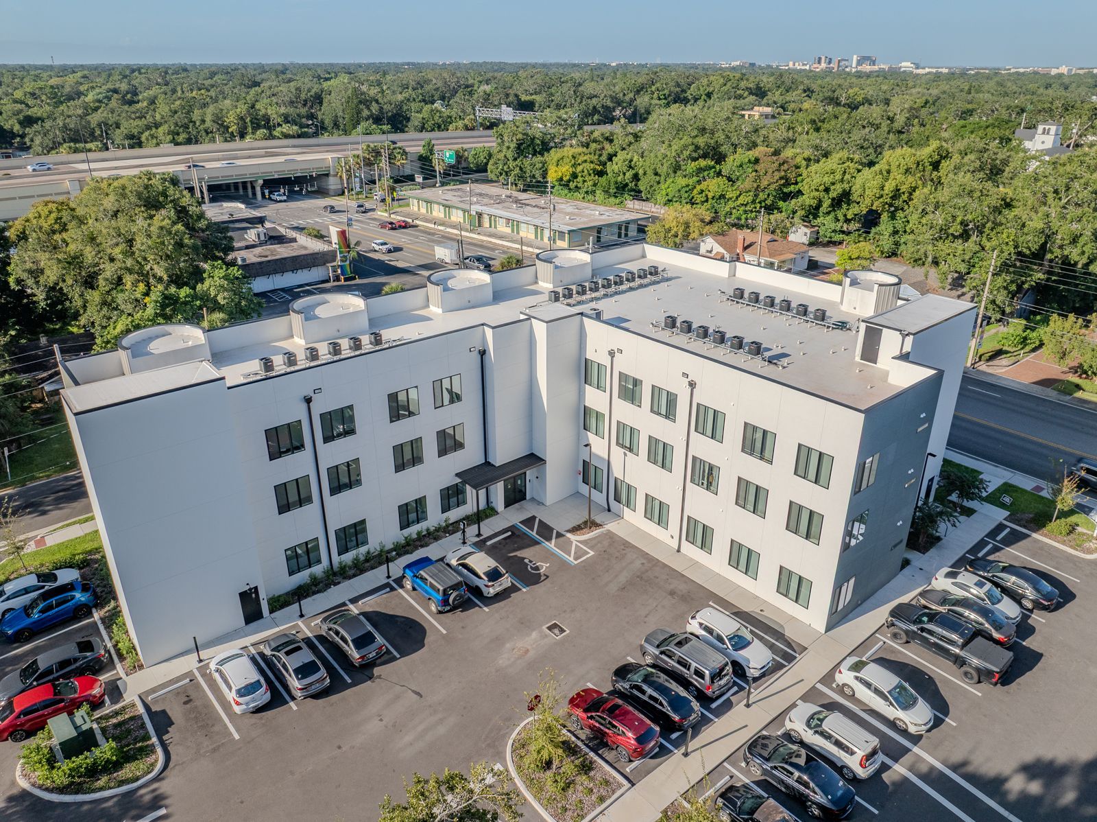 Aerial view of a modern white building with a parking lot filled with cars, surrounded by trees.