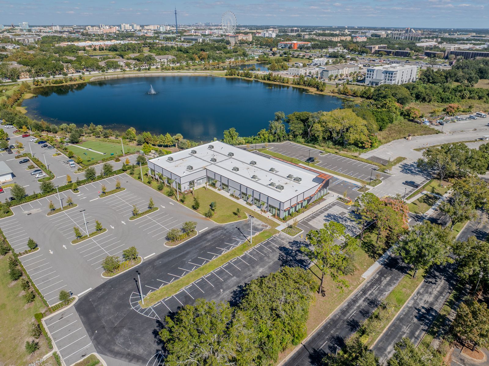 Aerial view: large building with parking lots, near a lake with surrounding trees and a city backdrop.