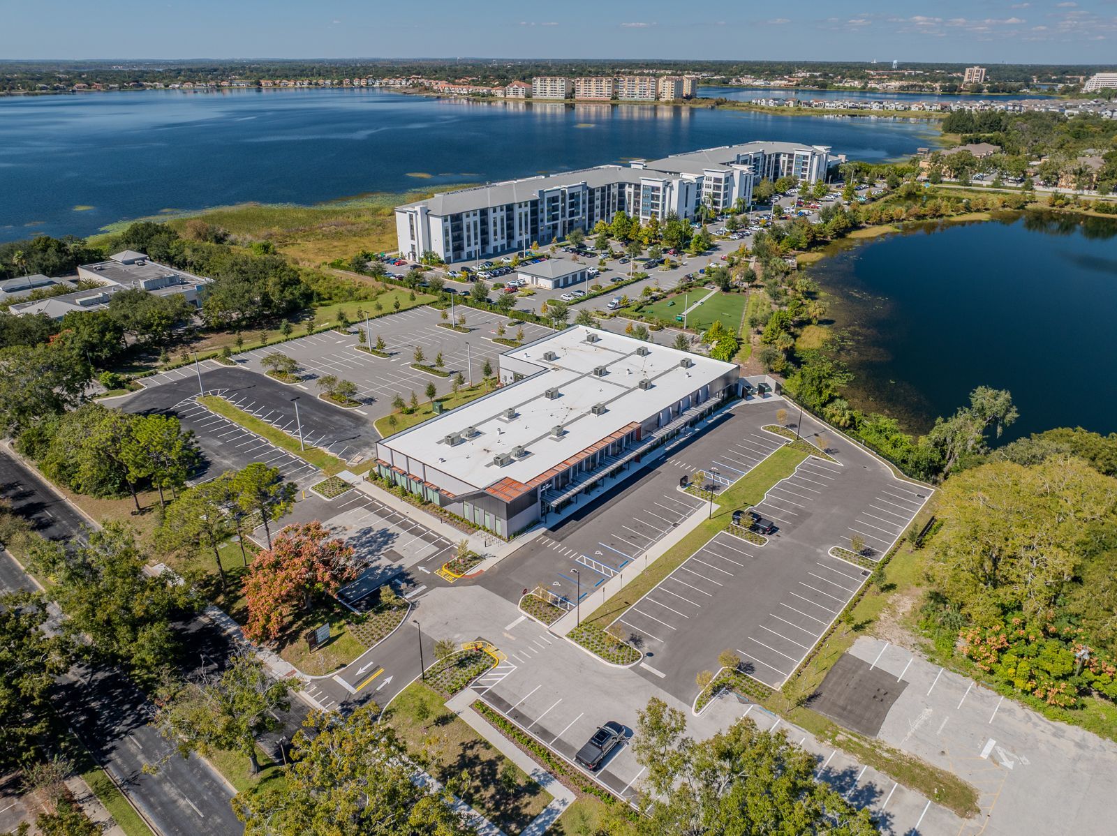 Aerial view of a modern building with large parking areas, situated near a lake with other buildings in the background.