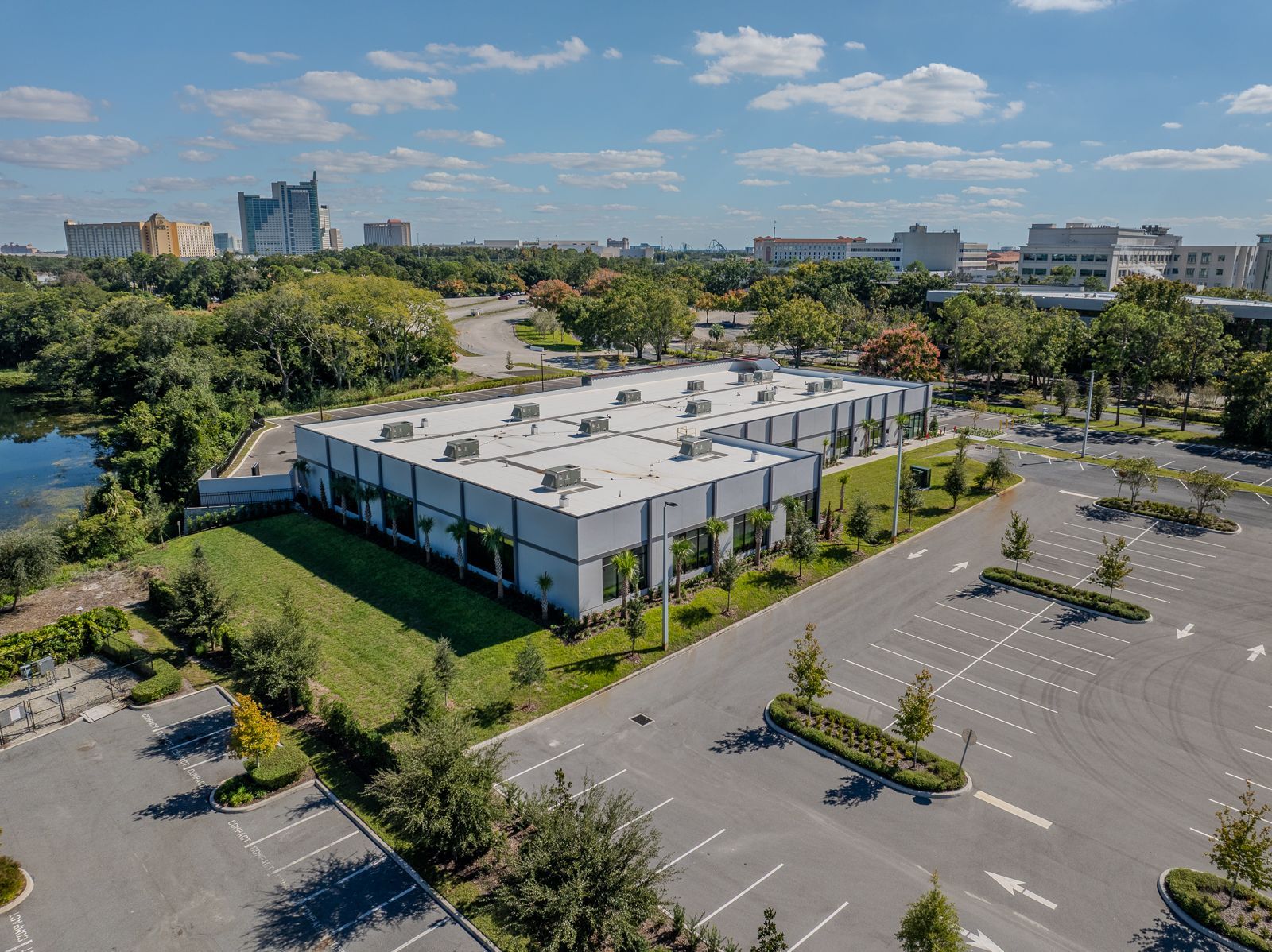 Aerial view of a modern gray building surrounded by a parking lot, trees, and a lake under a blue sky.