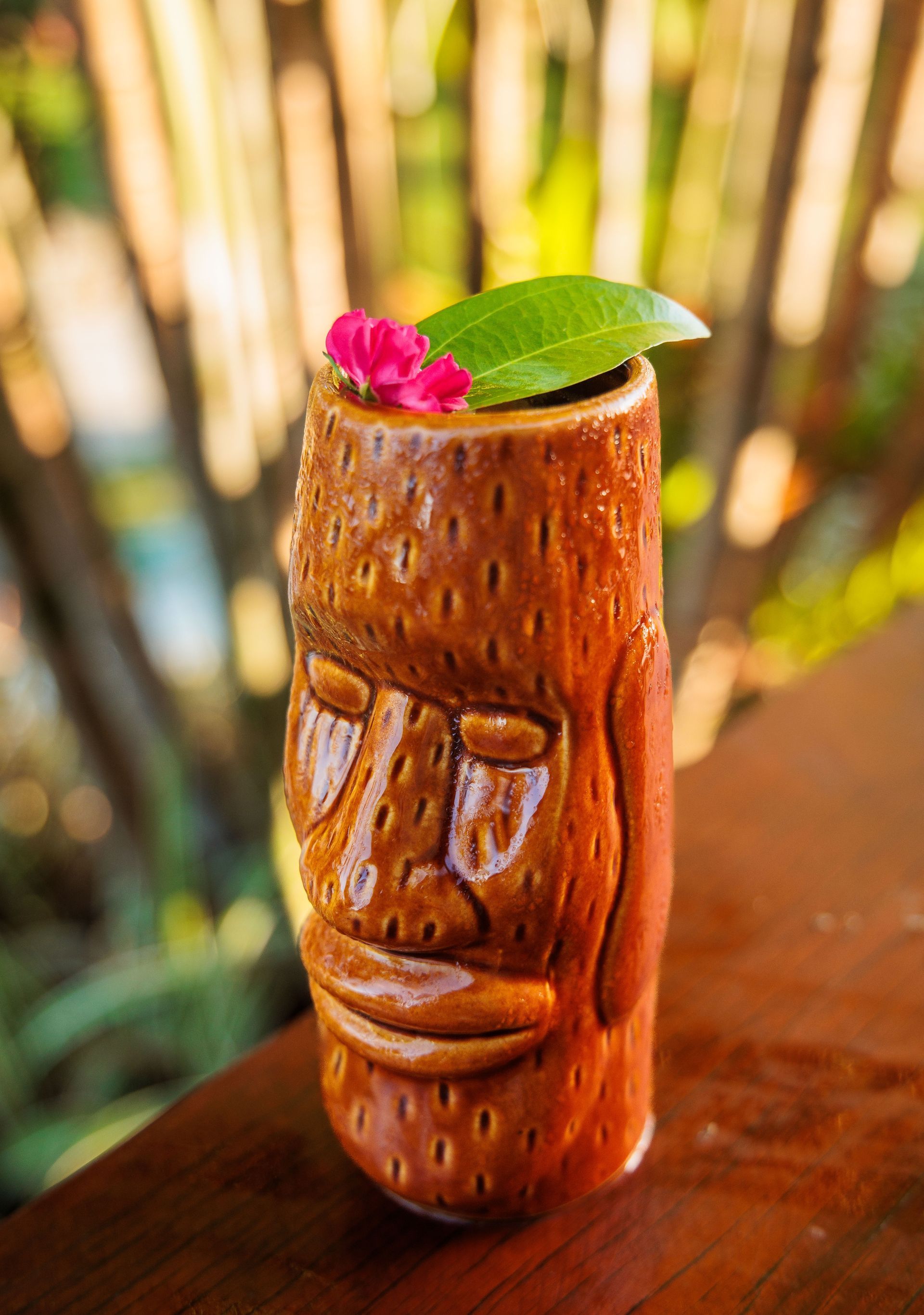 Uma caneca tiki com uma flor dentro está sobre uma mesa de madeira.