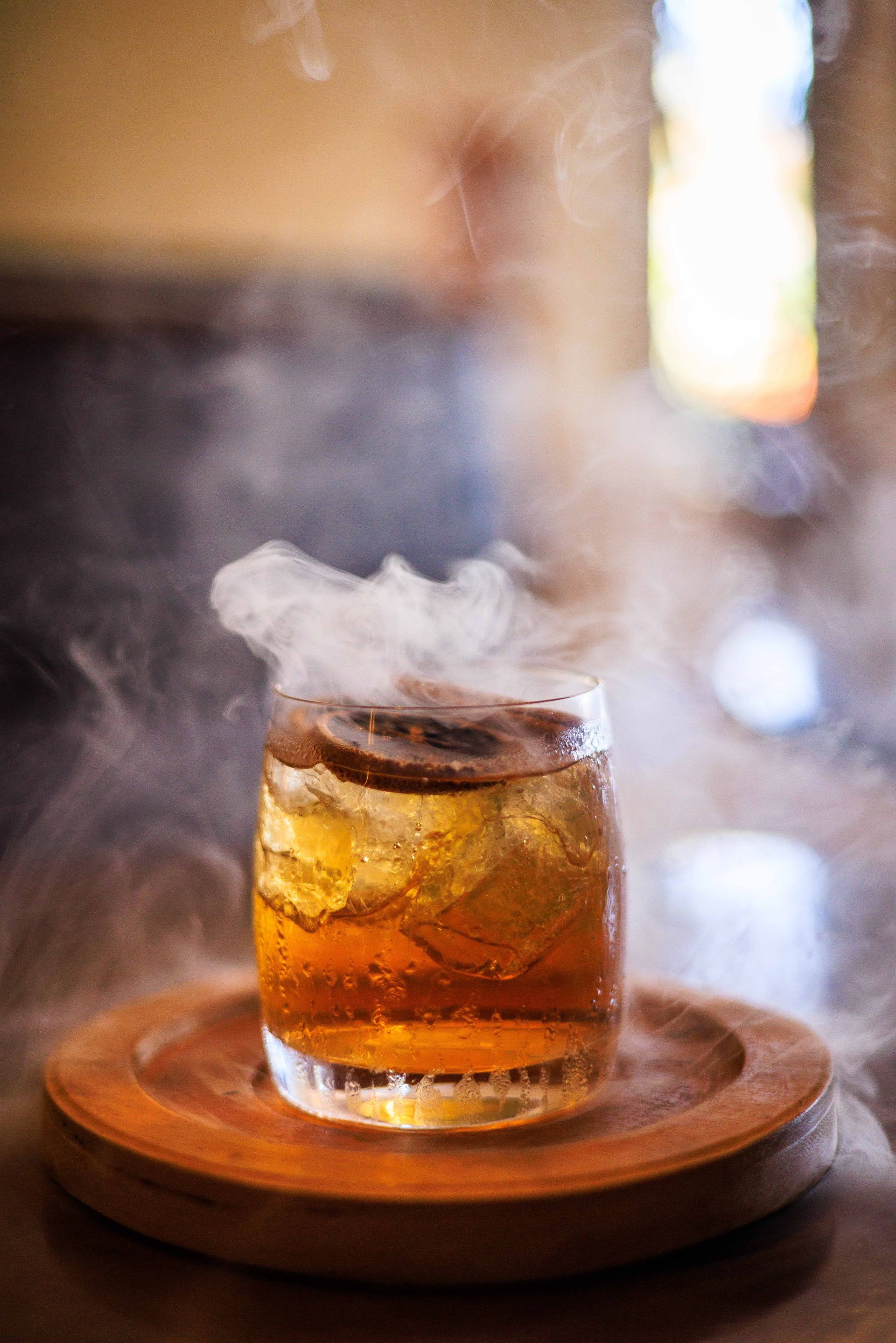 Um close de um copo de uísque com fumaça saindo dele em um prato de madeira.