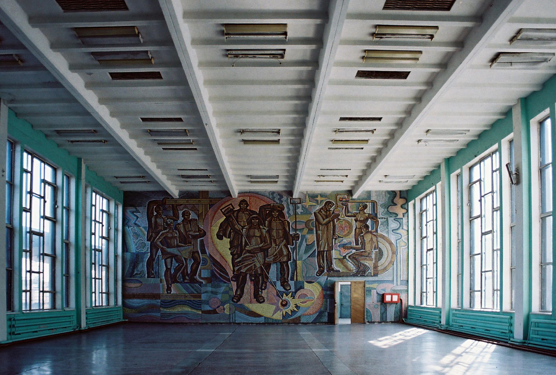 Large, empty room with teal walls, large windows, and a mural depicting figures; sunlight streams in.