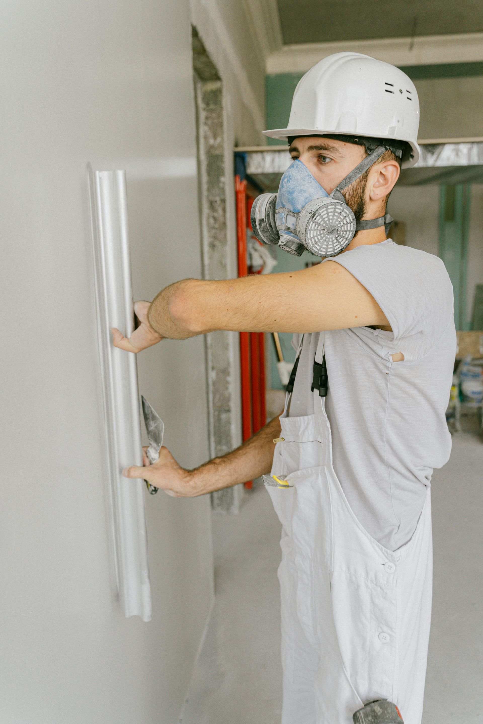 Construction worker in white overalls and hard hat smoothing drywall, wearing a respirator mask.