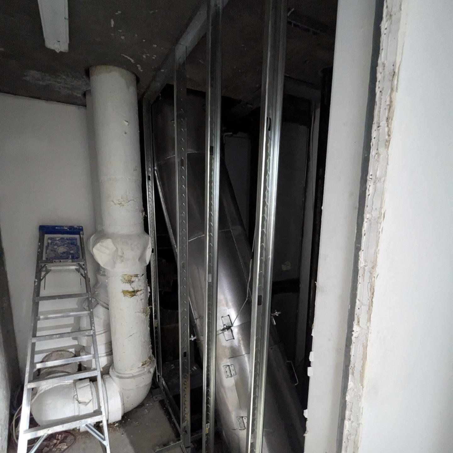 Construction in a room: metal framing, white pipes, ladder, and drywall opening.