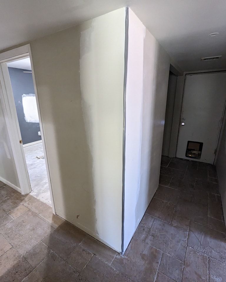 Hallway with a freshly painted wall in the center, leading to doors and rooms with beige floors.