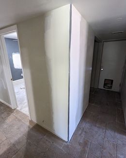 Hallway with a freshly painted wall in the center, leading to doors and rooms with beige floors.