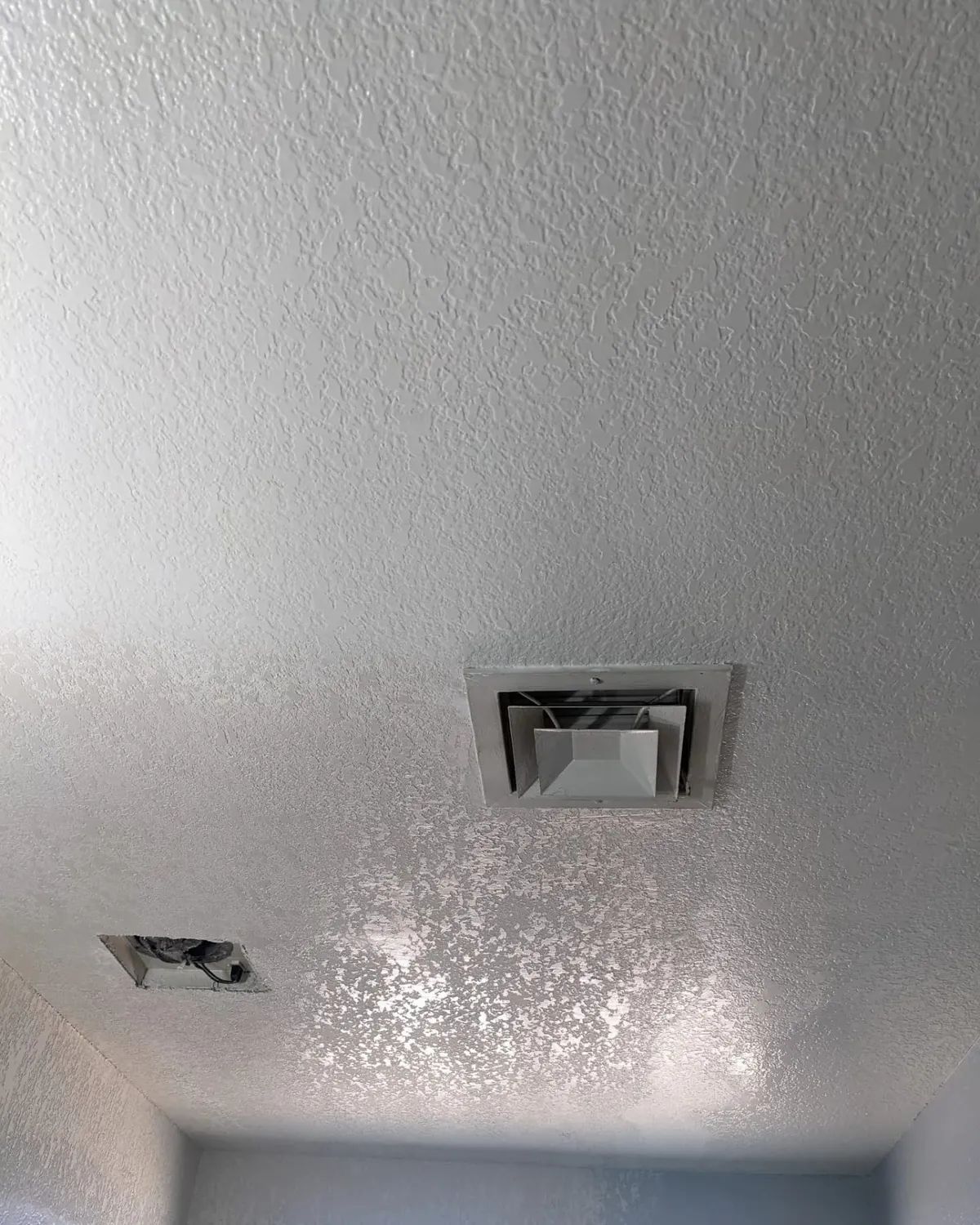 White textured ceiling with two square vents, the lower one surrounded by speckled residue.