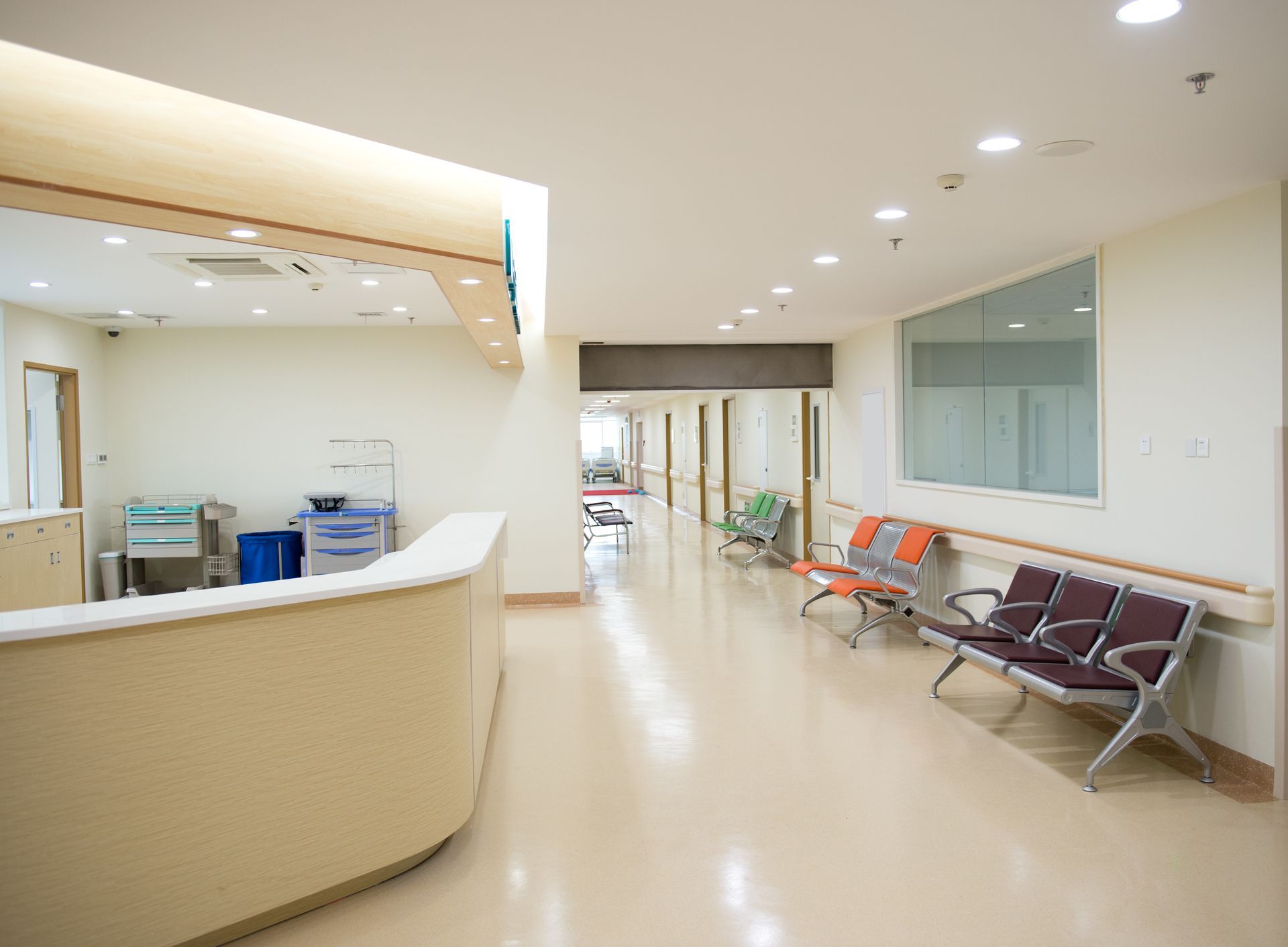 Hospital hallway with reception desk and waiting area. Beige and orange seating. Smooth, light-colored flooring.