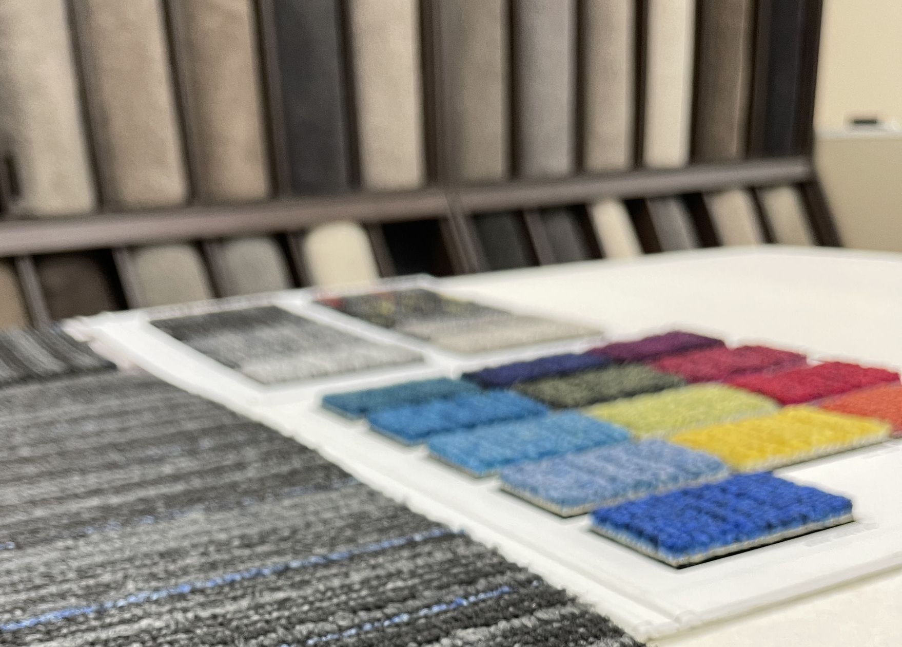 Carpet samples: gray, blue, and colorful swatches on a white surface, with a rack of carpet samples in the background.