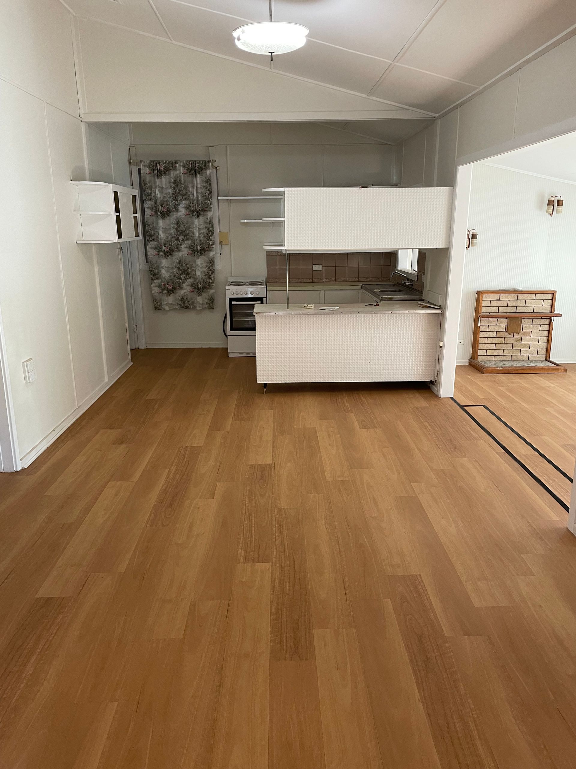 Interior of a small living space with a kitchen area. Wooden floors, white walls, and a light fixture.