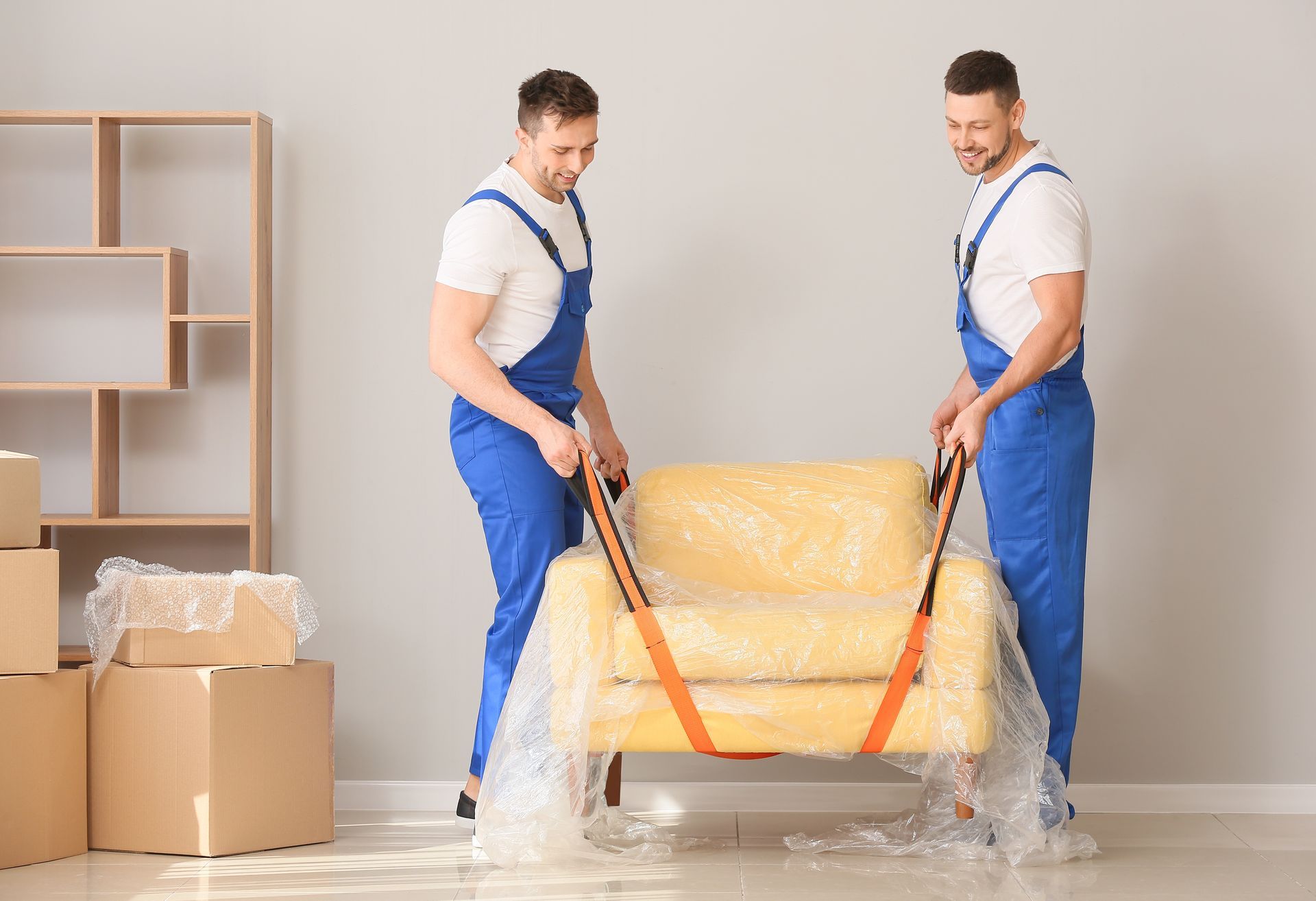 Two movers carry a yellow sofa with moving straps in a flat with cardboard boxes and a bookstand. Two movers carry a yellow sofa with moving straps in a flat with cardboard boxes and a bookstand.