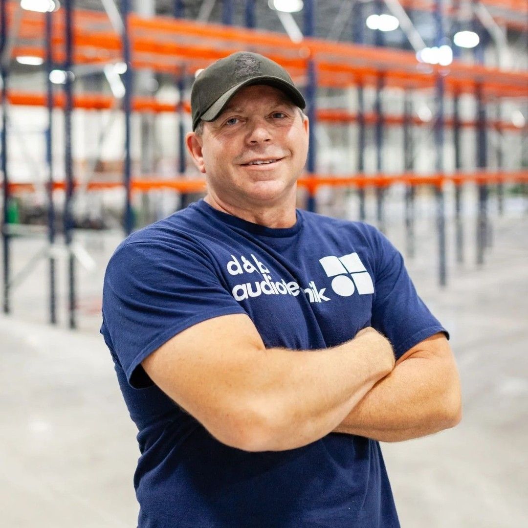 Man in blue shirt and baseball cap with arms crossed, in warehouse.