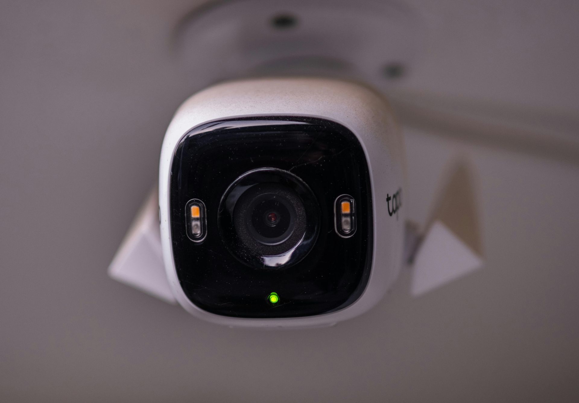 Security camera mounted on a white ceiling, featuring a black lens, green light, and white housing.