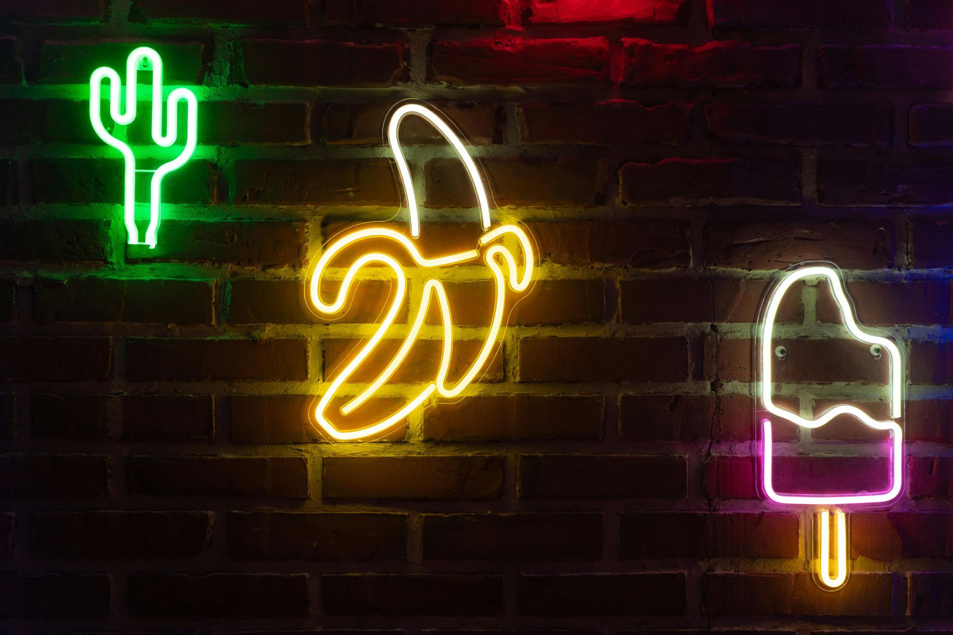Neon signs of a green cactus, yellow peeled banana, and pink popsicle on a brick wall.