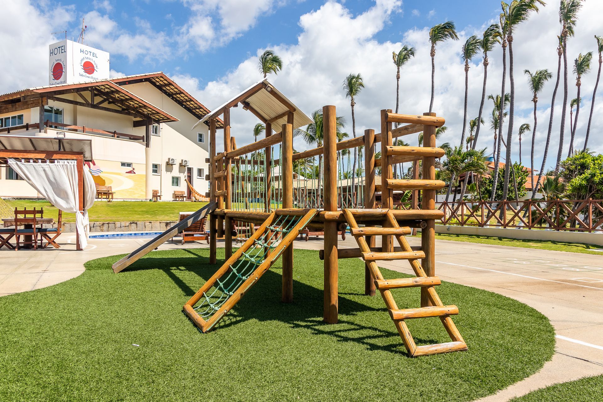 Wooden playground on artificial grass next to a building and pool.