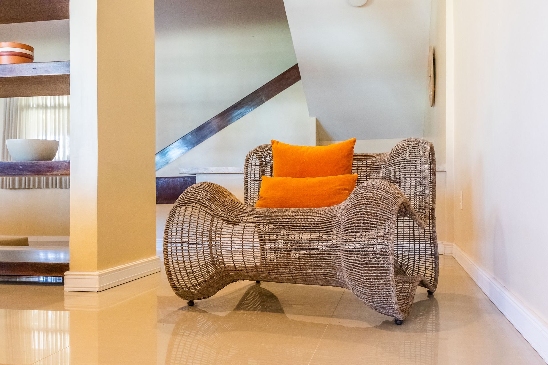 Wicker armchair with orange pillows in a sunlit entryway. Staircase and built-in shelf in the background.