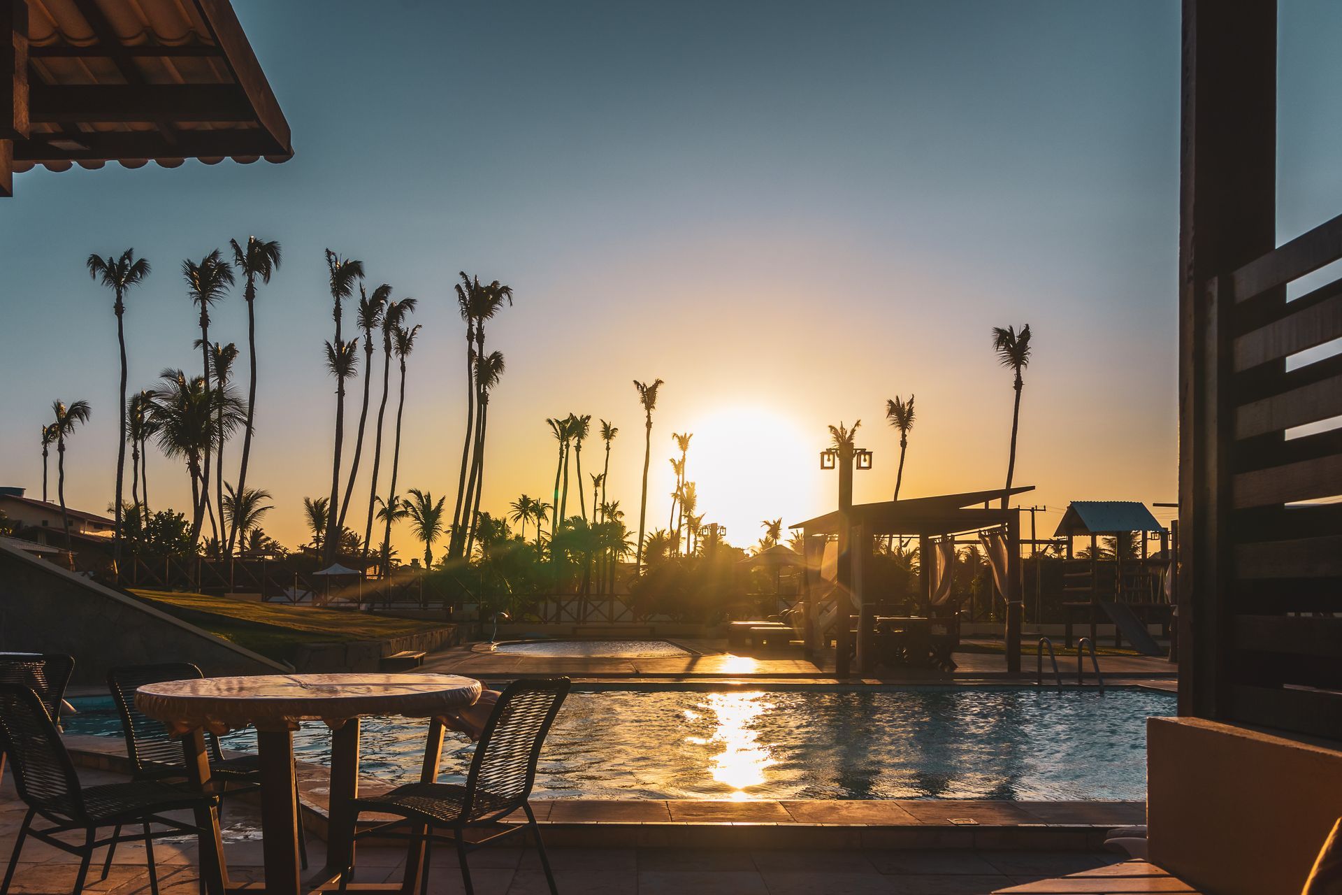Sunset view over a pool, palm trees silhouetted against a golden sky, with tables and chairs in the foreground.