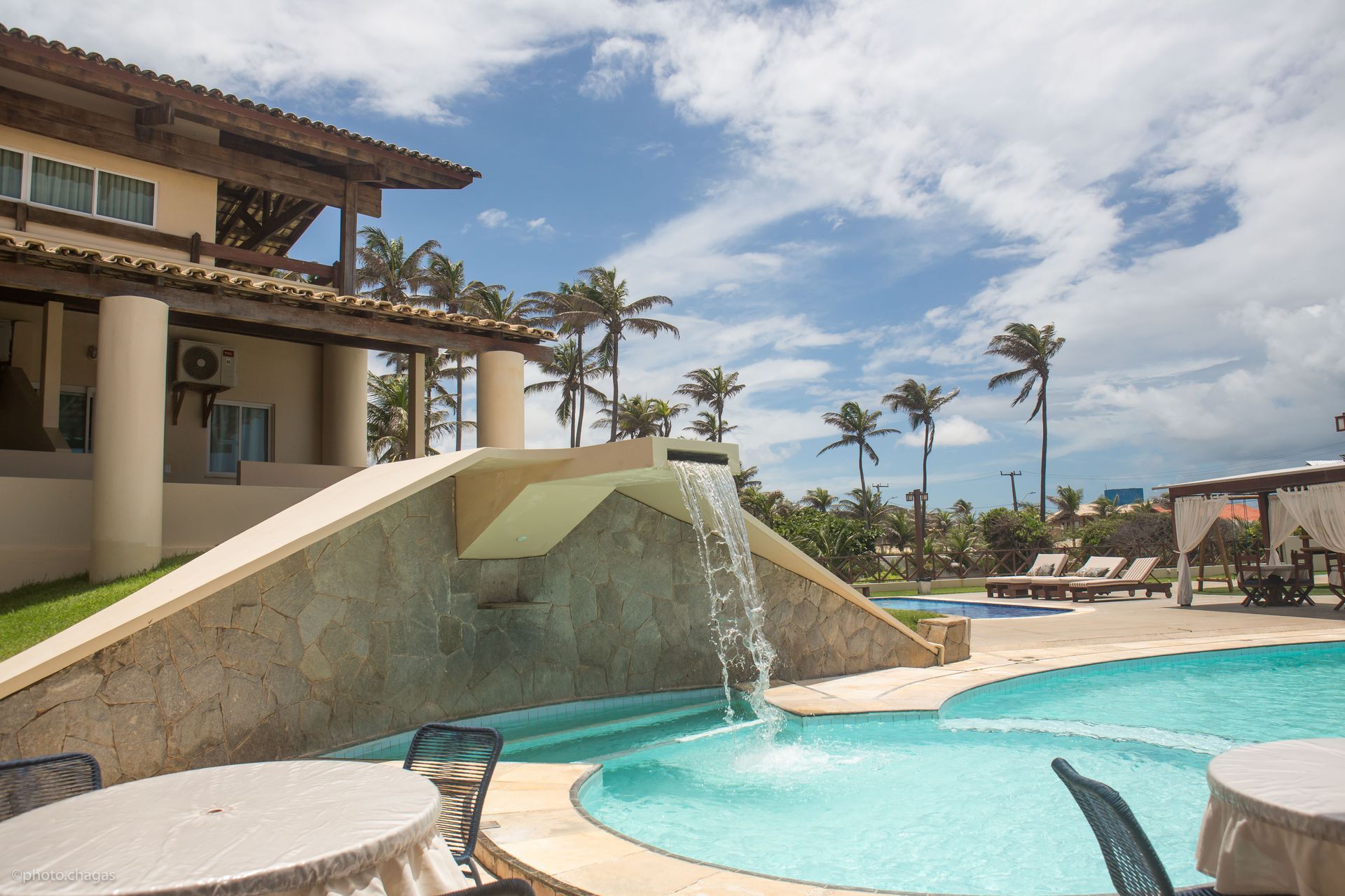 Luxurious resort with a pool, waterfall, and palm trees under a blue sky.